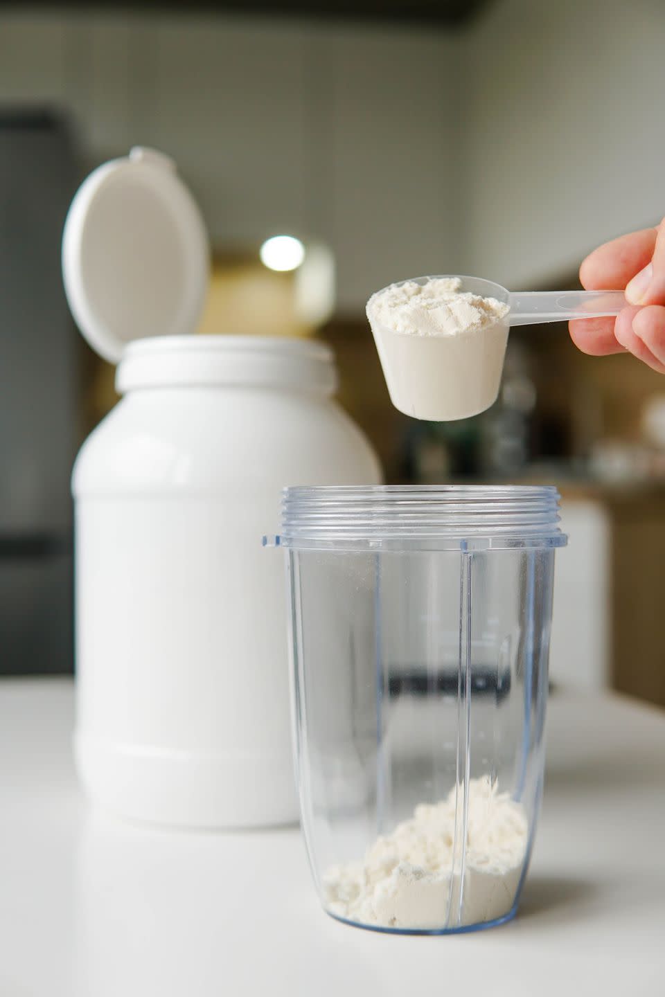 young man with measuring spoon puts portion of whey protein powder into a shaker on wooden table, making protein drink