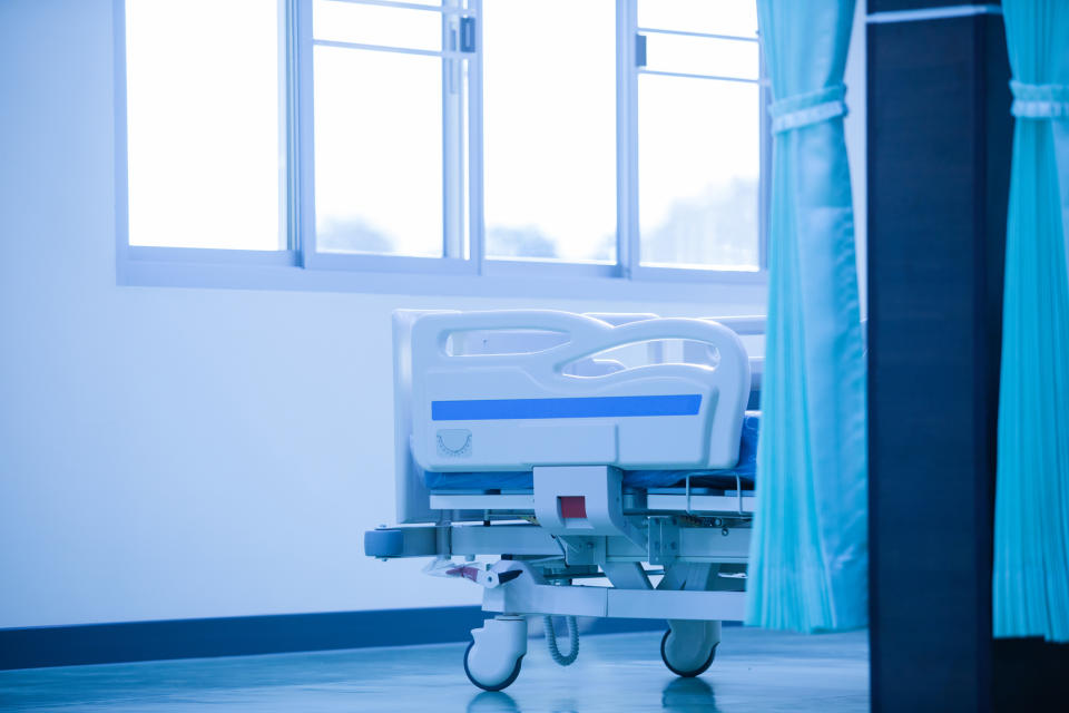 A hospital bed with adjustable height and attached equipment is positioned near large windows with light blue curtains partially visible