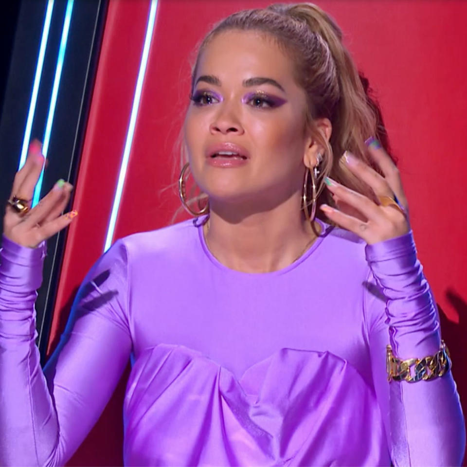 Rita Ora dressed in purple, with her hair tied up and gold hoop earrings, is visibly upset while sitting in her red The Voice chair.