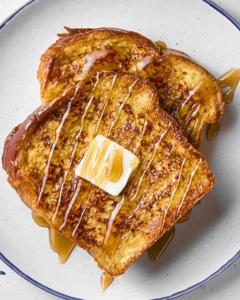 overhead shot of two pieces of french toast on a white plate with a blue rim, topped with a pat of butter and drizzled with maple syrup