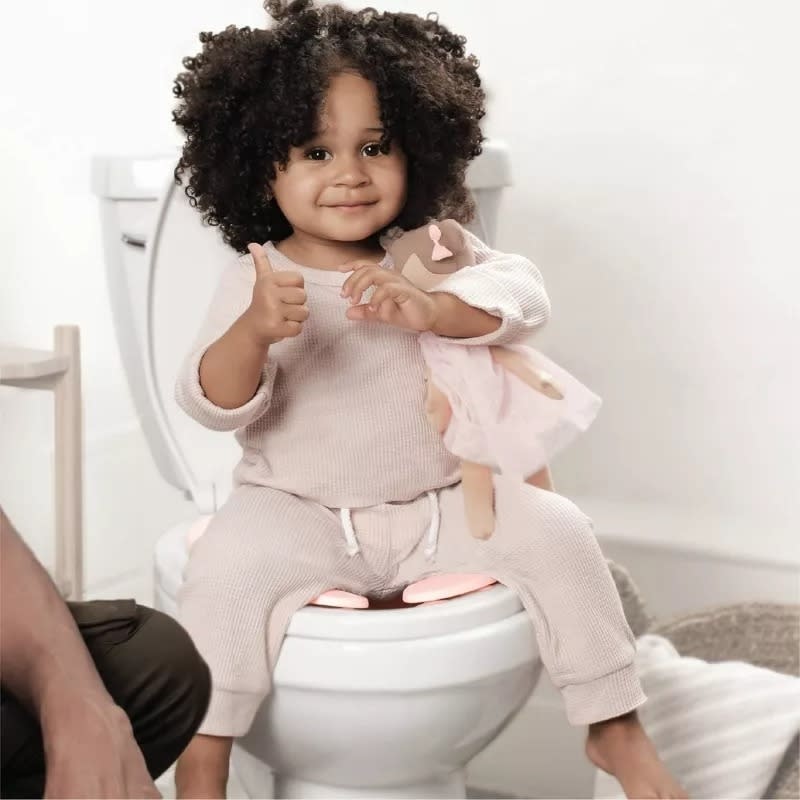 Child on a potty training seat, holding a doll, giving a thumbs up, with an adult's leg nearby