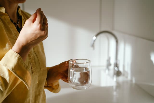 <p>Fiordaliso / Getty Images</p> A female in a yellow shirt with a glass of water and taking vitamins.