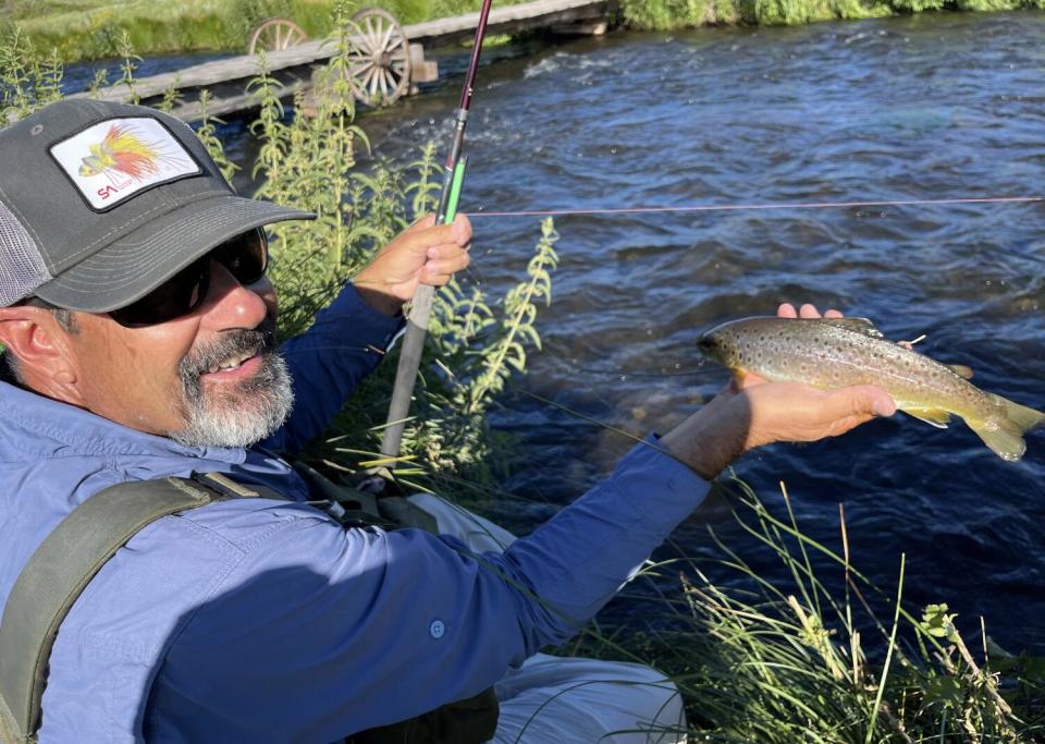 A man holds a fishing pole and a rainbow trout beside a river.