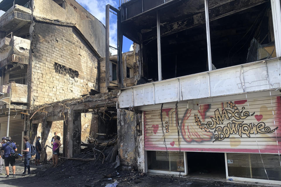 Reporters work by charred shop and building in a s street of Pointe-a-Pitre, Guadeloupe island, Sunday, Nov.21, 2021. French authorities are sending police special forces to the Caribbean island of Guadeloupe, an overseas territory of France, as protests over COVID-19 restrictions erupted into rioting. In Pointe-a-Pitre, the island's largest urban area, clashes left three people injured, including a 80-year-old woman who was hit by a bullet while on her balcony. A firefighter and a police officer were also injured and several shops were looted there and in other towns. (AP Photo/Elodie Soupama)