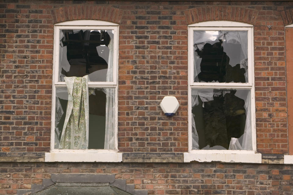 Two windows of a brick building with broken glass panes and torn curtains visible