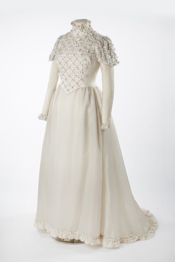 wedding dress made by neymar, worn by sara raiher for her wedding on january 25th 1972 full length dress with a line skirt, full length sleeves with puffed shoulders and high neckline bodice is decorated with cross hatched pattern of embellishment, beads and fabric flowers same flowers appear on cuffs and hem