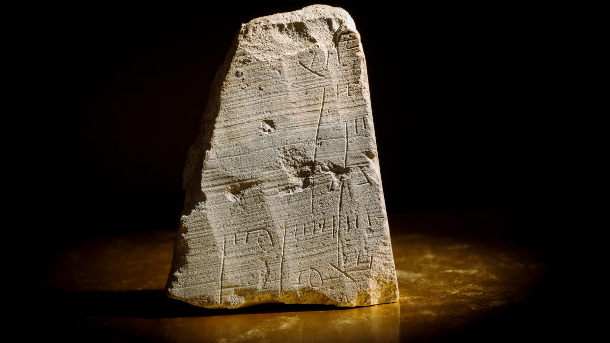  We see a triangular gray stone slab sitting that is inscribed with Hebrew against a black background. 