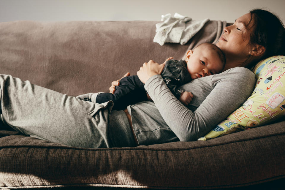 A woman is lying on a couch with her eyes closed, cradling a baby who is also lying peacefully on her chest