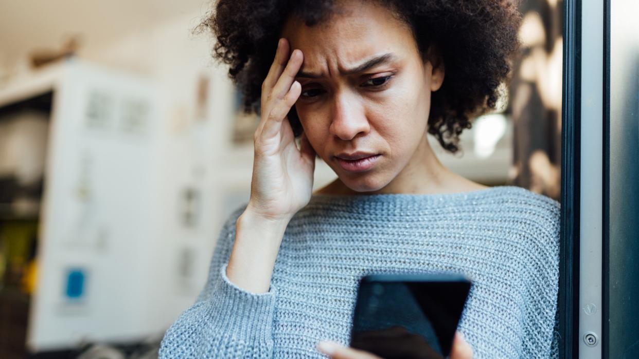  Woman looks anxiously at her phone with one hand on her forehead . 