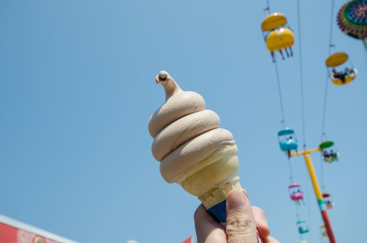 hand holds up a chocolate soft serve ice cream against the bright blue sky with amusement park ride in background