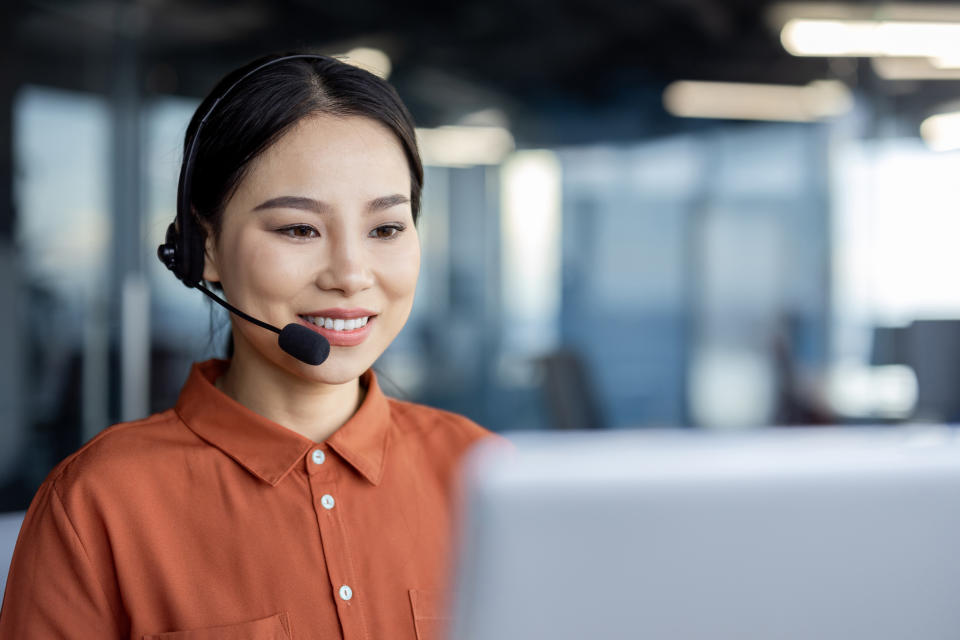 A woman with a headset and microphone is smiling while looking at her computer screen. She appears to be in a modern office setting