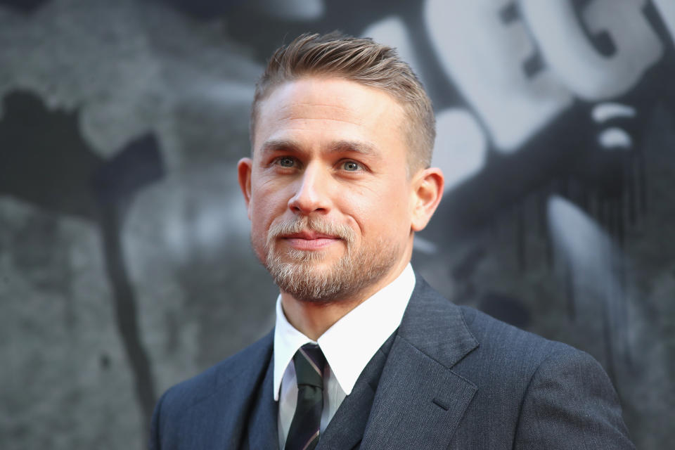 Charlie Hunnam smiling on the red carpet, wearing a dark suit with a tie and vest