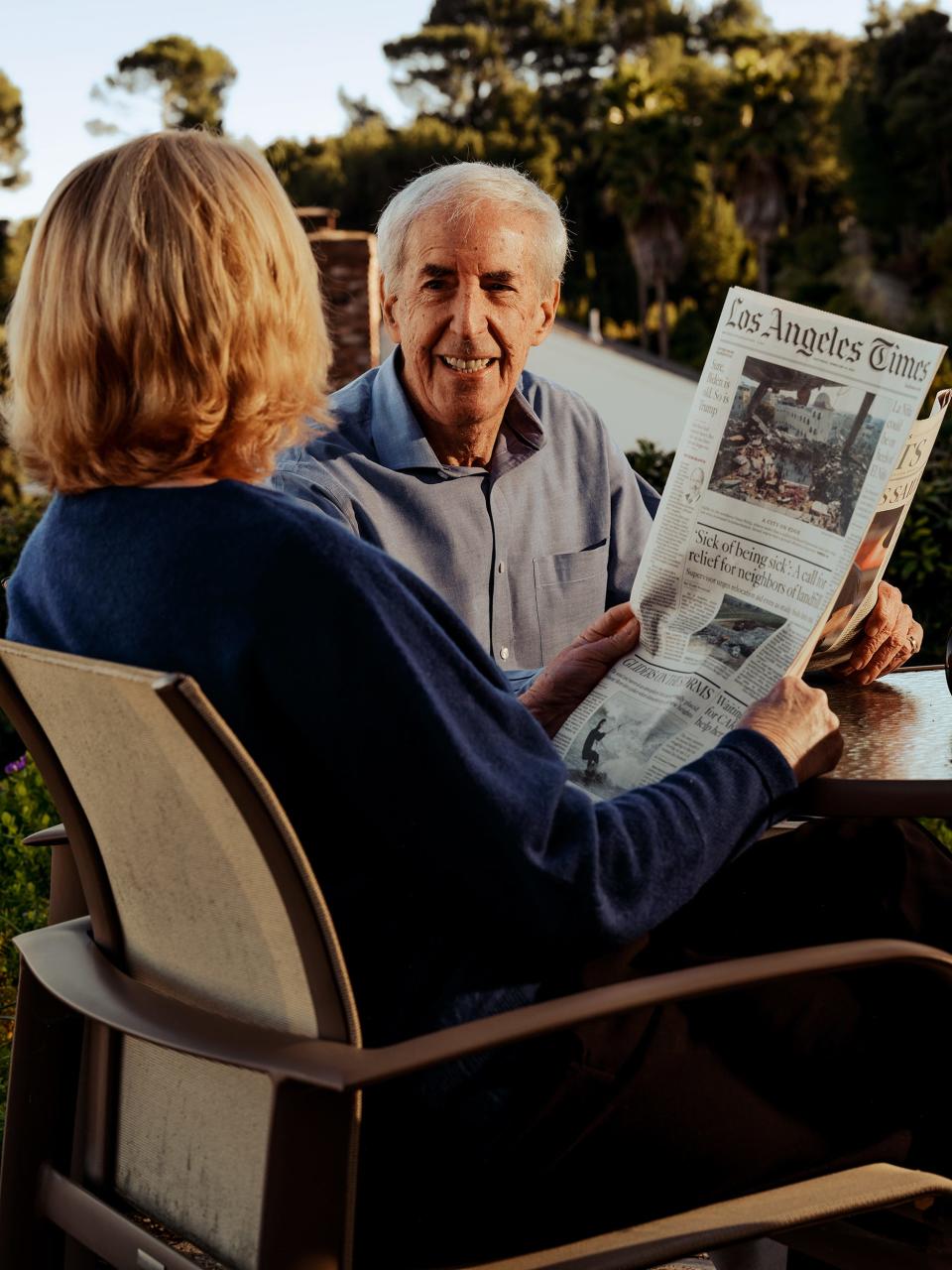 Man smiling at his wife as they both sit outside reading newspapers