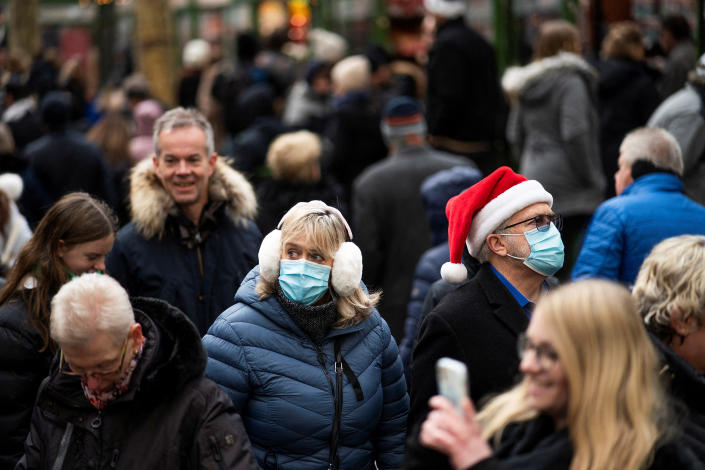 Several dozen people in winter weather clothes stand or walk outside, some wear face masks.