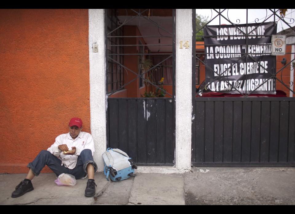 In this May 17, 2012 photo, Krambilm Kennedy, a migrant from Nicaragua, leans up against a wall and eats, on the opposite side of the street of a shelter for migrants, where a sign hangs from a wrought iron fence that reads in Spanish: "The neighbors of Lecheria demand the closure of the migrant house," in Lecheria, on the outskirts of Mexico City. While the number of Mexicans heading to the U.S. has dropped dramatically, a surge of Central American migrants is making the 1,000-mile northbound journey this year, fueled in large part by the rising violence brought by the spread of Mexican drug cartels. (AP Photo/Alexandre Meneghini)