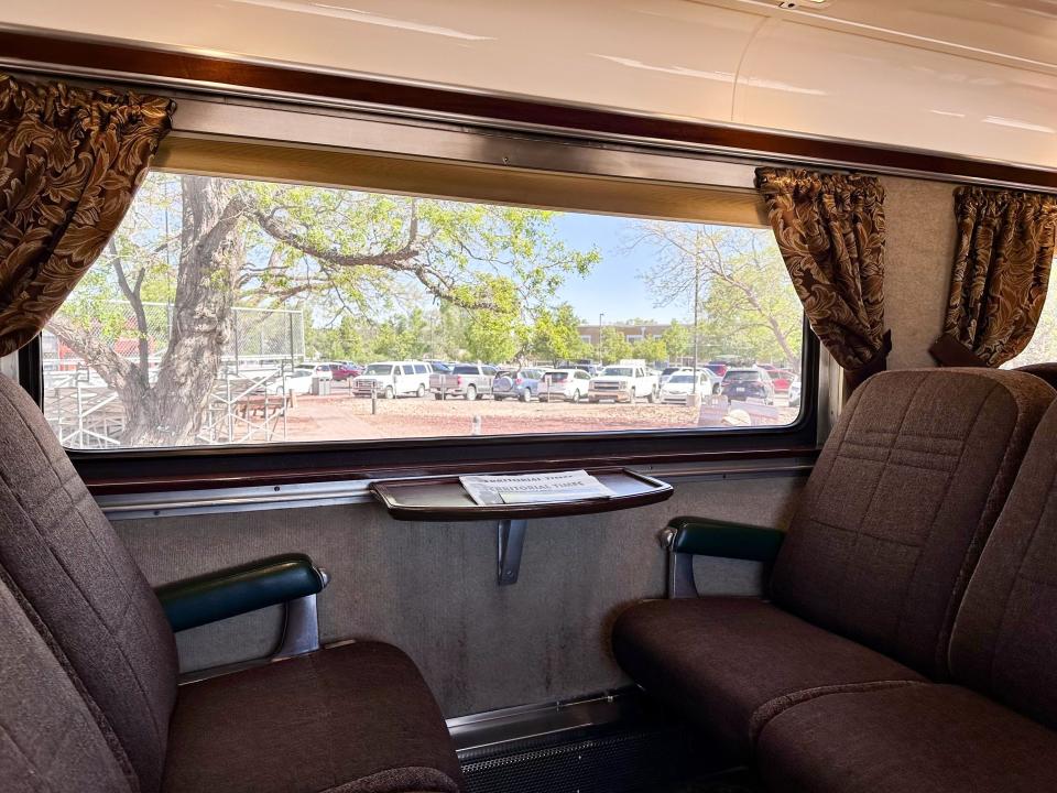 The first-class car on the Grand Canyon Railway, which includes two sets of two plush brown seats facing each other, a small table with a newspaper, a large window, and brown patterned curtains.