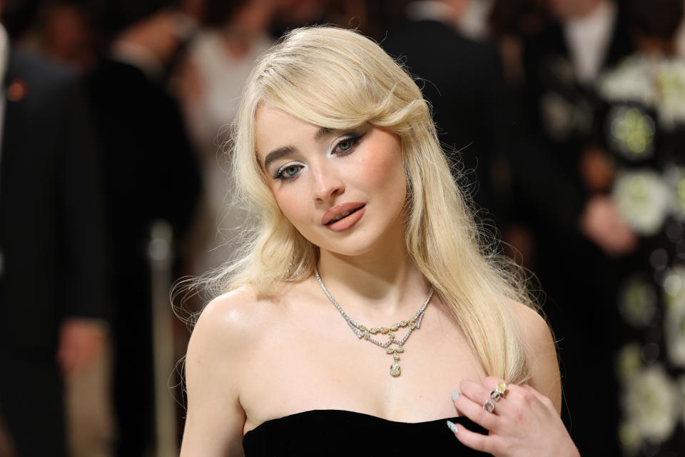 Sabrina Carpenter with long, blonde hair and dark strapless attire poses, wearing a necklace and ring, with a blurred crowd in the background