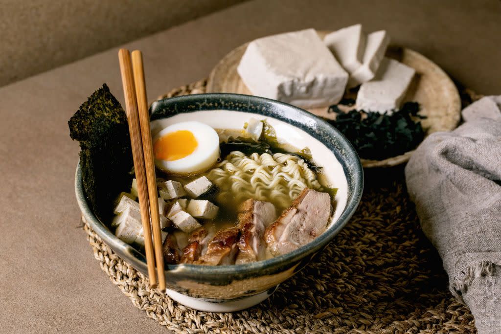 homemade japanese style soup ramen with noodles, grilled duck breast, tofu, seaweed nori chips and boiled egg in ceramic bowl with chopsticks and spoon on brown table with knitted straw napkin