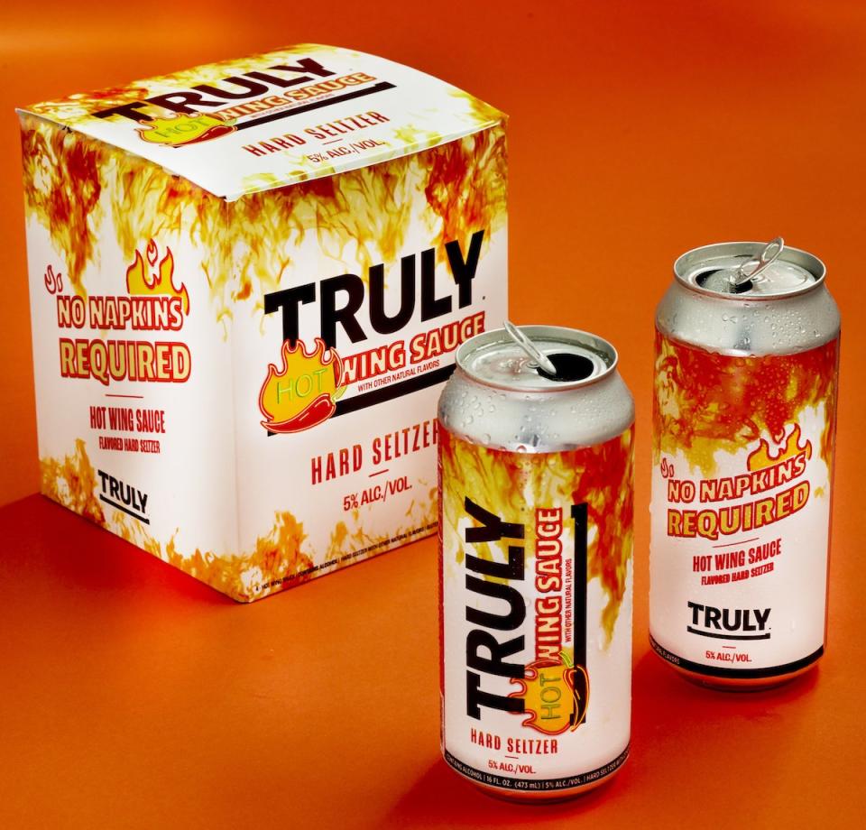 A white and orange/red box of Truly Hot Wing Sauce Hard Seltzer with two opens can in front of it