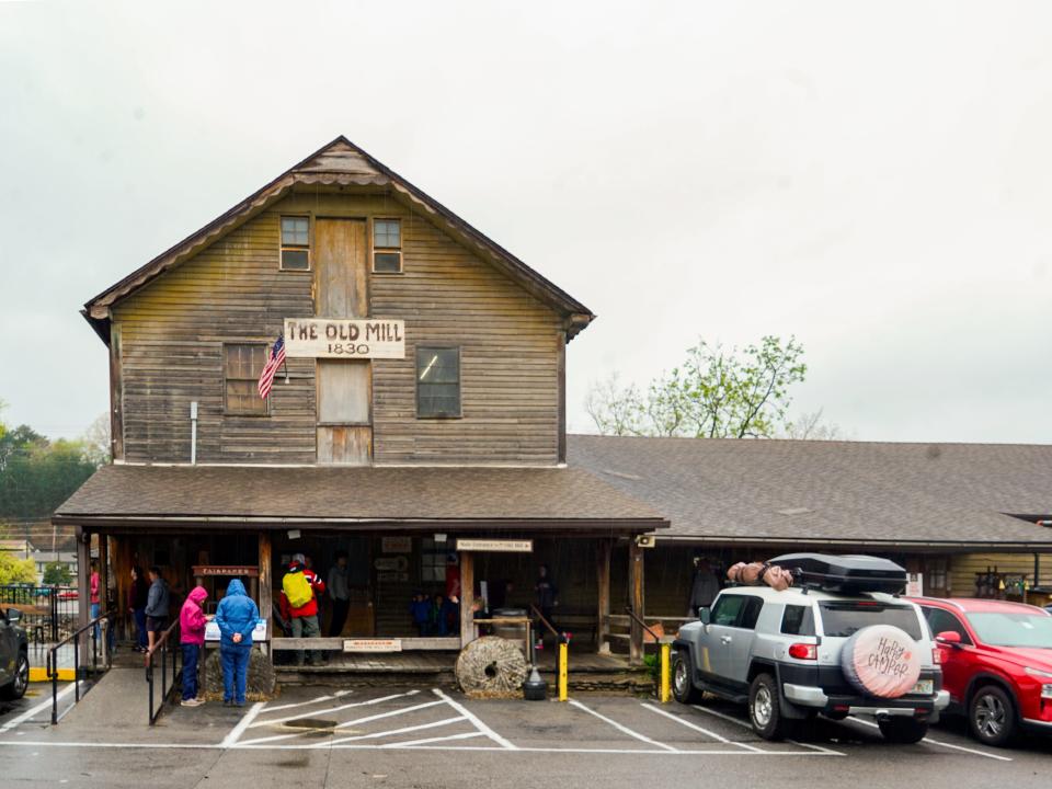 An old wooden building with a triangular roof behind a parking lot full of cars. Skies are gray behind it.