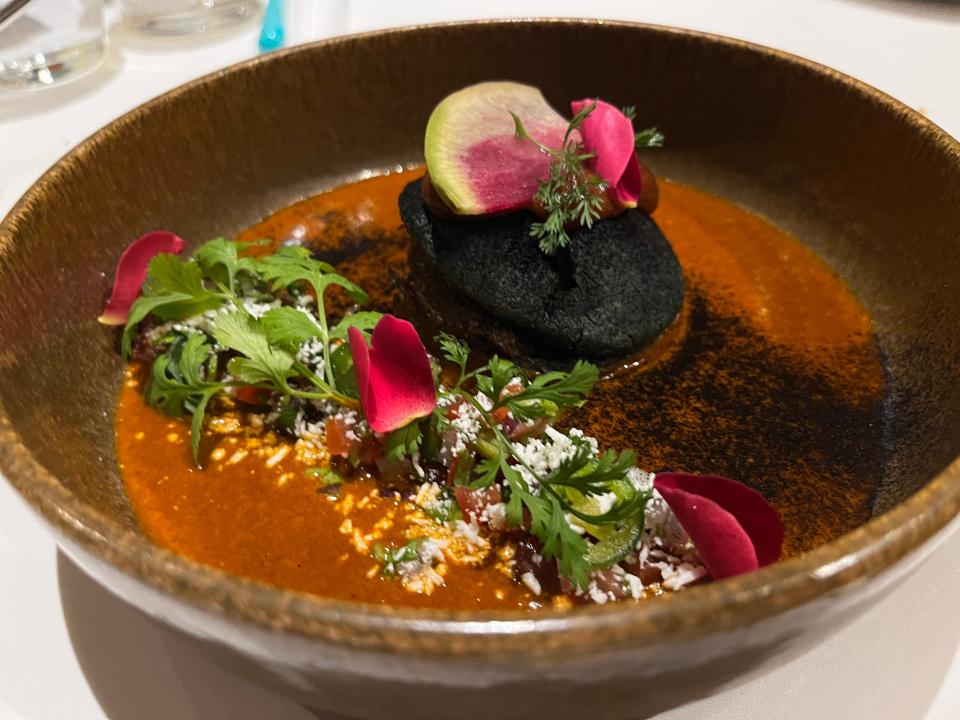 A bowl of red soup with herbs and edible flowers on top. The soup is in a brown bowl.