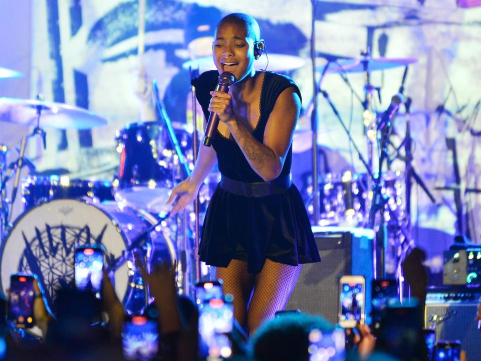 WILLOW performs on stage, backlit with blue light, in front of a sea of recording cell phones. She's wearing a short black skirt, fishnets, and a black fitted tee with a sweetheart neckline.