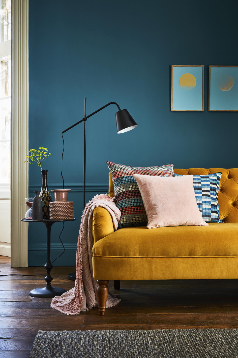turquoise blue living room with mustard velvet couch, hardwood floor, graphic print cushions, pink blanket, black floor lamp, side table with vases