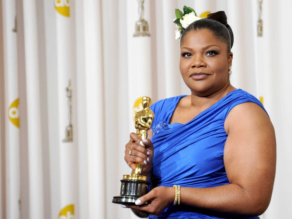 Mo'nique holding an Oscar statue in front of a rippled white background while wearing a blue sleeveless dress.