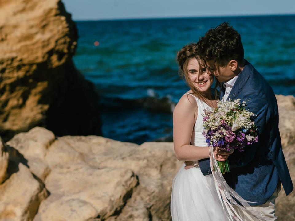 a bride and groom holding flowers on rocks by a beach