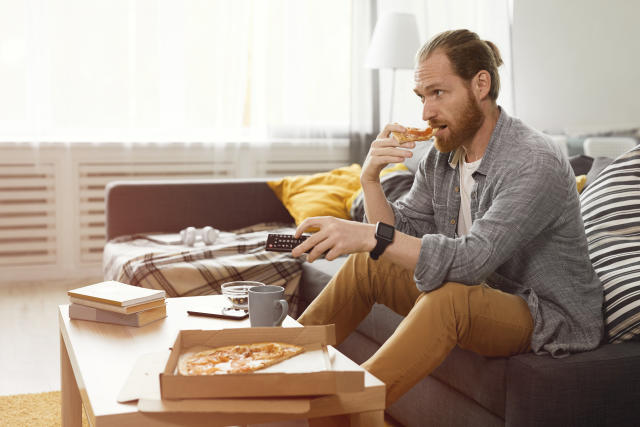 Side view portrait of bearded man eating pizza while watching TV at home in bachelors pad, copy space