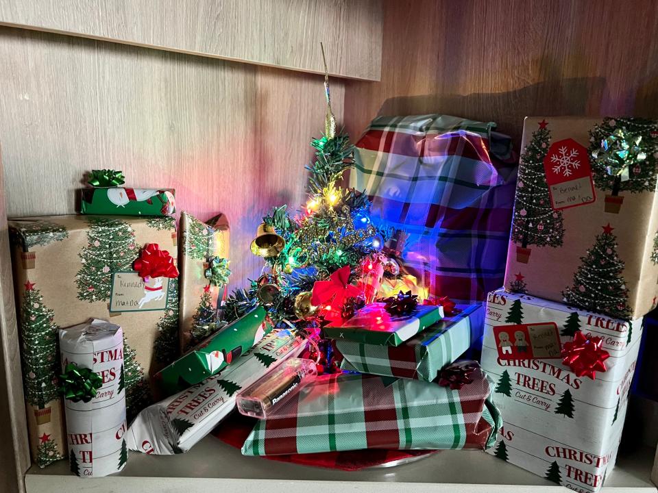 Image of a tiny Christmas tree that has gold bells, ornaments, a red bow, and lights. It's surrounded by wrapped Christmas presents and sits on a wooden shelf.