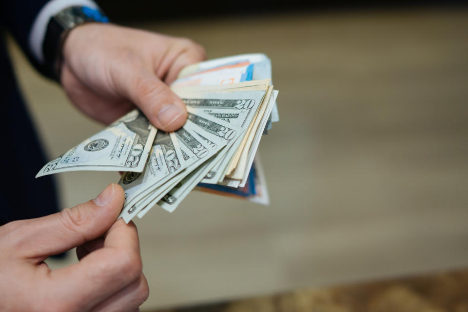 Hands holding a fanned-out stack of U.S. dollar bills, including twenties, fifties, and hundreds, as well as various other banknotes