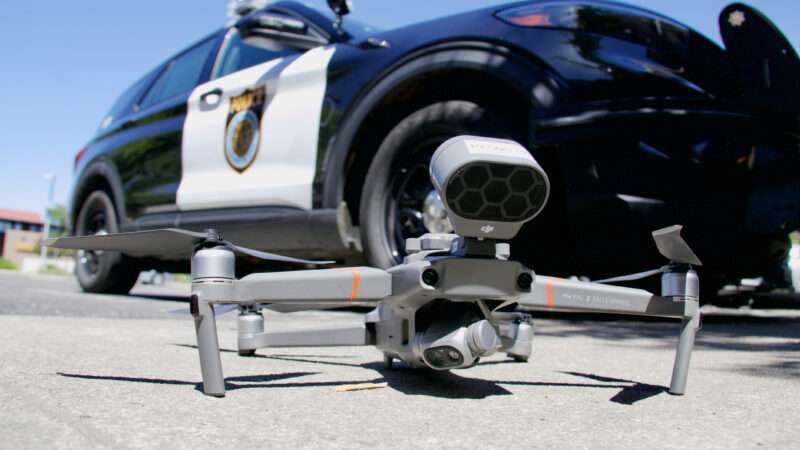 A drone with a camera attached sits on the ground, with a police car in the background.