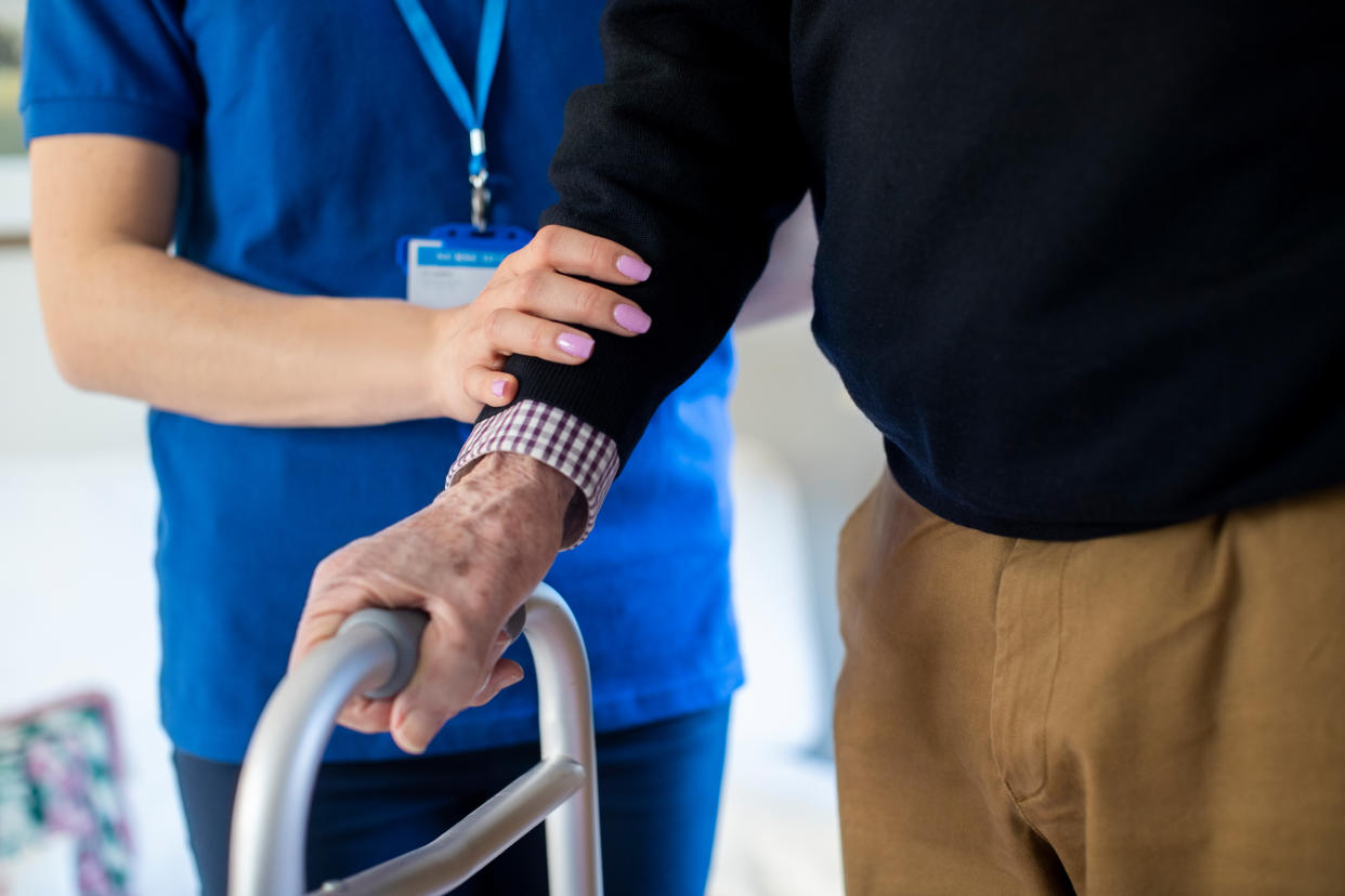 Close Up Of Senior Man With Hands On Walking Frame Being Helped By Care Worker