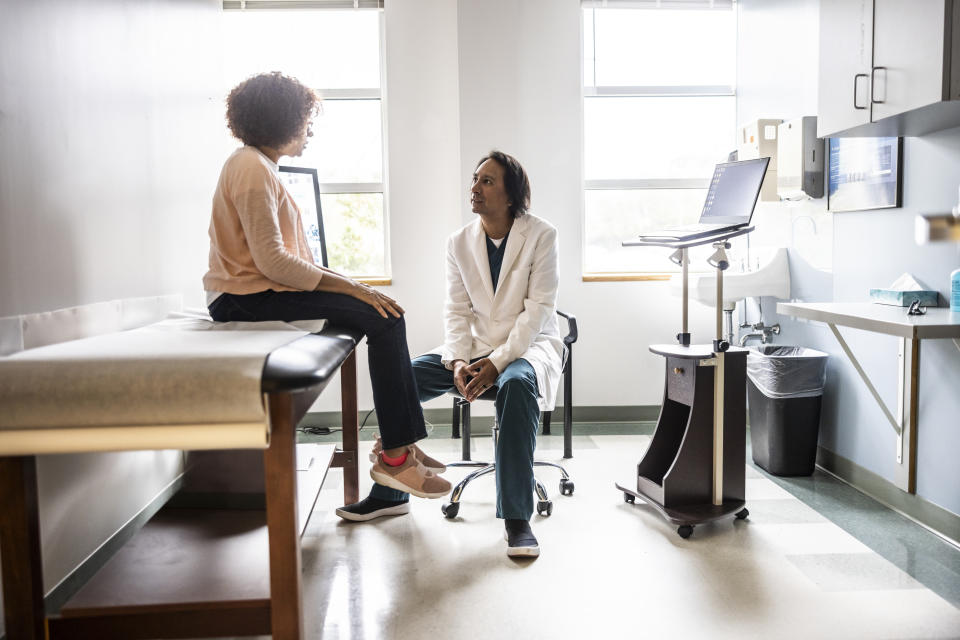 A patient sits on an examination table talking with a doctor in a white coat in a medical consultation room