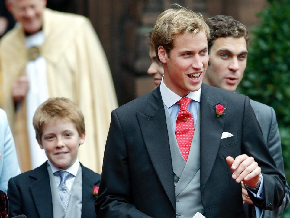 Hugh Grosvenor smiling while wearing a black suit with a grey tie and walking with Prince William ahead of a wedding group.