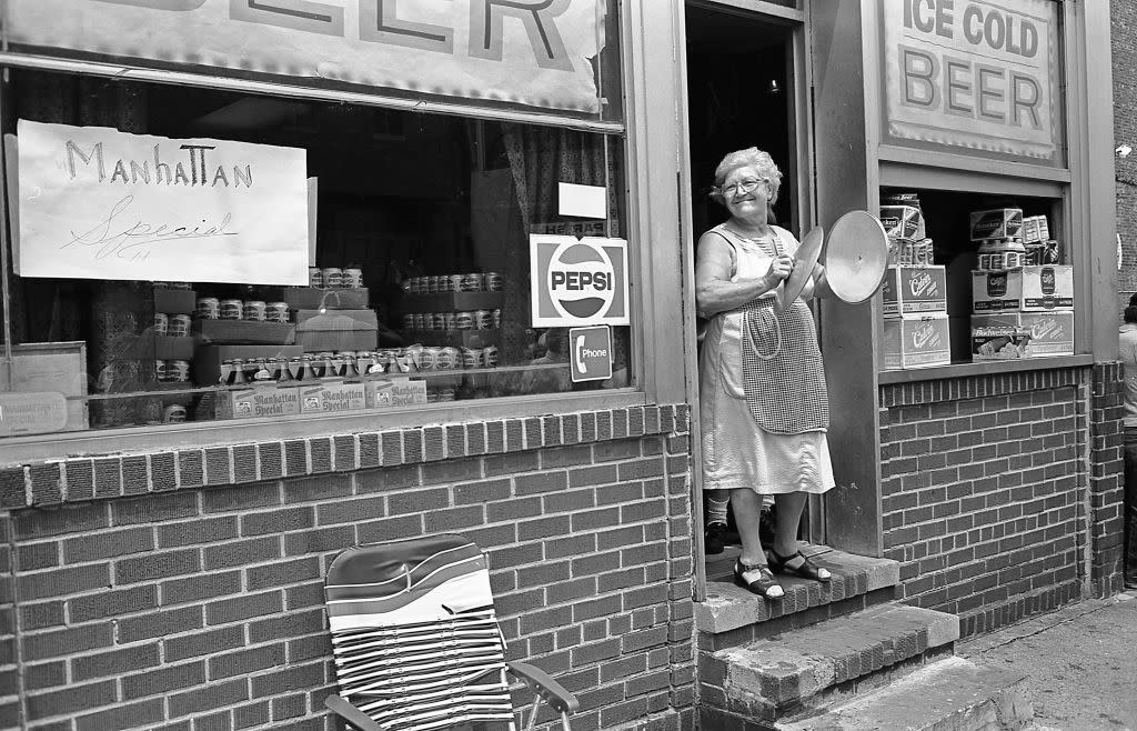 An elderly woman smiles as she claps the lids of two pots together In the open doorway of a shop