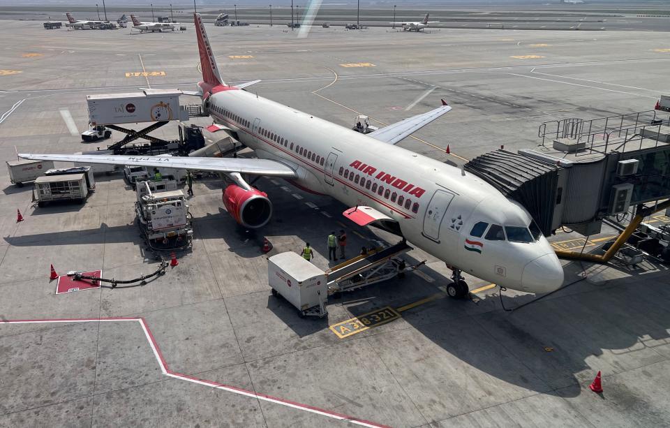 A white plane with red accents and "Air India" written on it waits at an airport, with a skybridge attached and workers loading luggage.