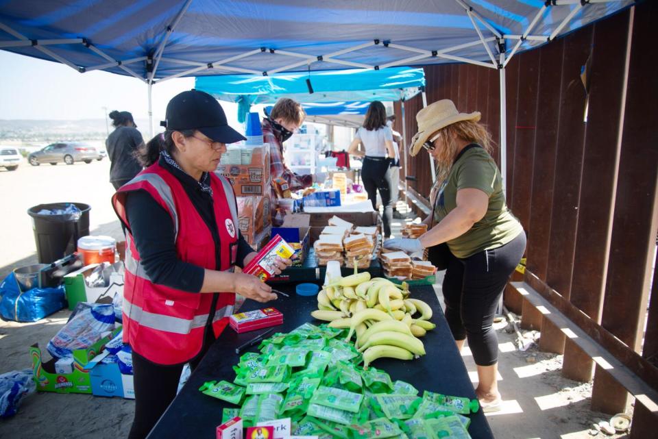 People, one in a red safety vest, near a table filled with bananas and other items.