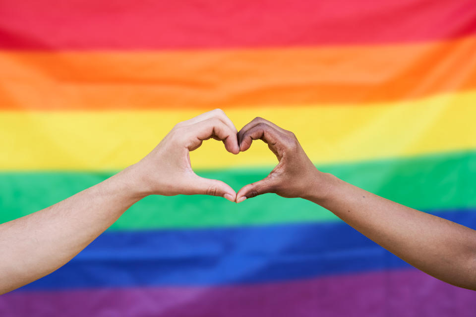 Heart shape made out of two diverse ethnicity hands with a background of a gay pride flag. White and black hands.