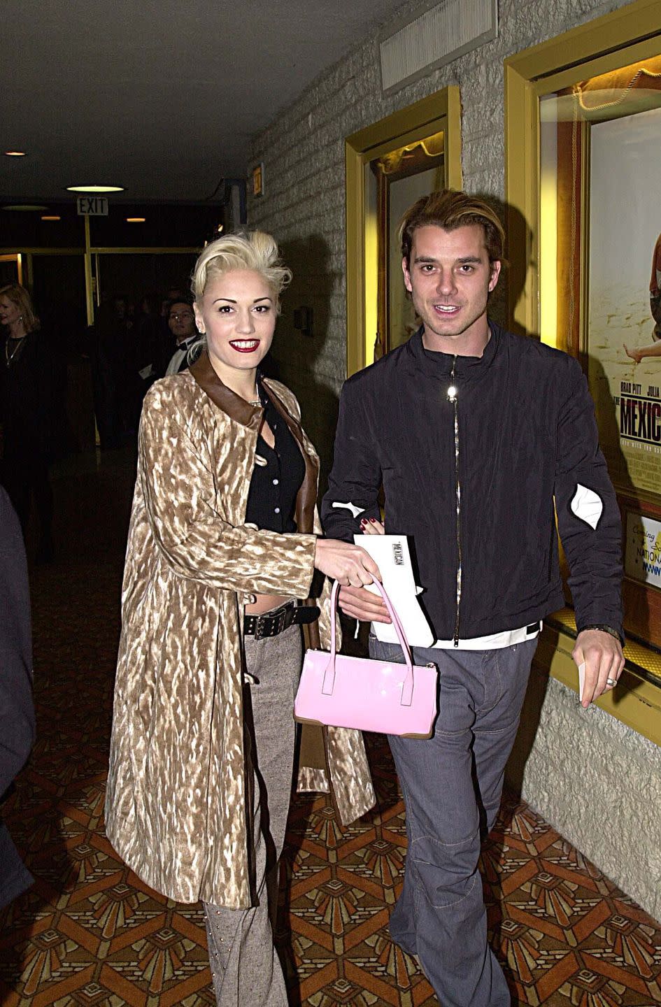 gwen stefani and gavin rossdale walk together next to a wall with movie posters on it, she wears a brown patterned coat and holds a pink purse, he wears a black jacket