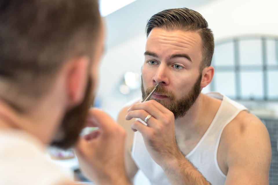 Handsome bearded young man standing plucking his nasal hairs in front of the mirror in the bathroom during his daily grooming