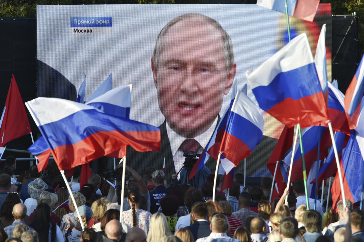 A screen shows President Vladimir Putin delivering his speech as people with red, white and blue flags stand watching.