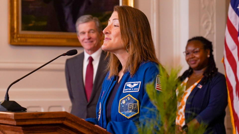 Eine Frau in einem blauen Fliegeranzug steht auf einem Podium mit zwei lächelnden Menschen und der amerikanischen Flagge hinter ihr.