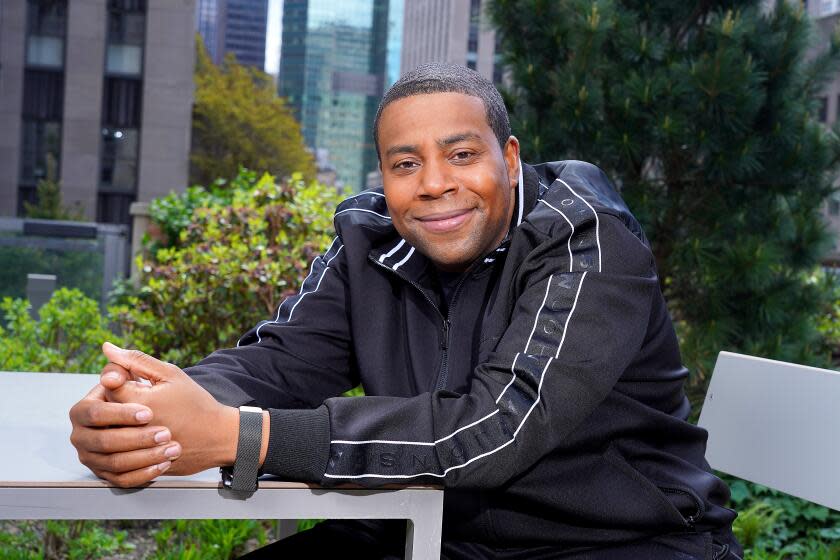 Kenan Thompson in a black jacket sitting on a metal chair with his hands clasped on a table