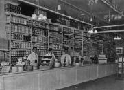 <p>Three grocery shop assistants stand behind the counter and in front of a tall shelf full of canned goods, as they wait for the next customer. </p>