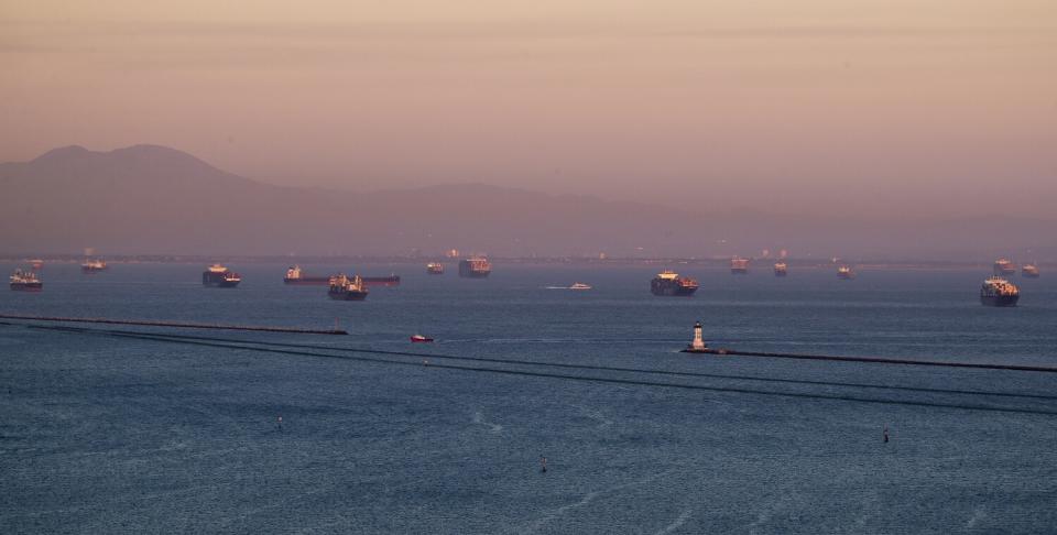 The sunset illuminates dozens of container ships in the ocean off the coast