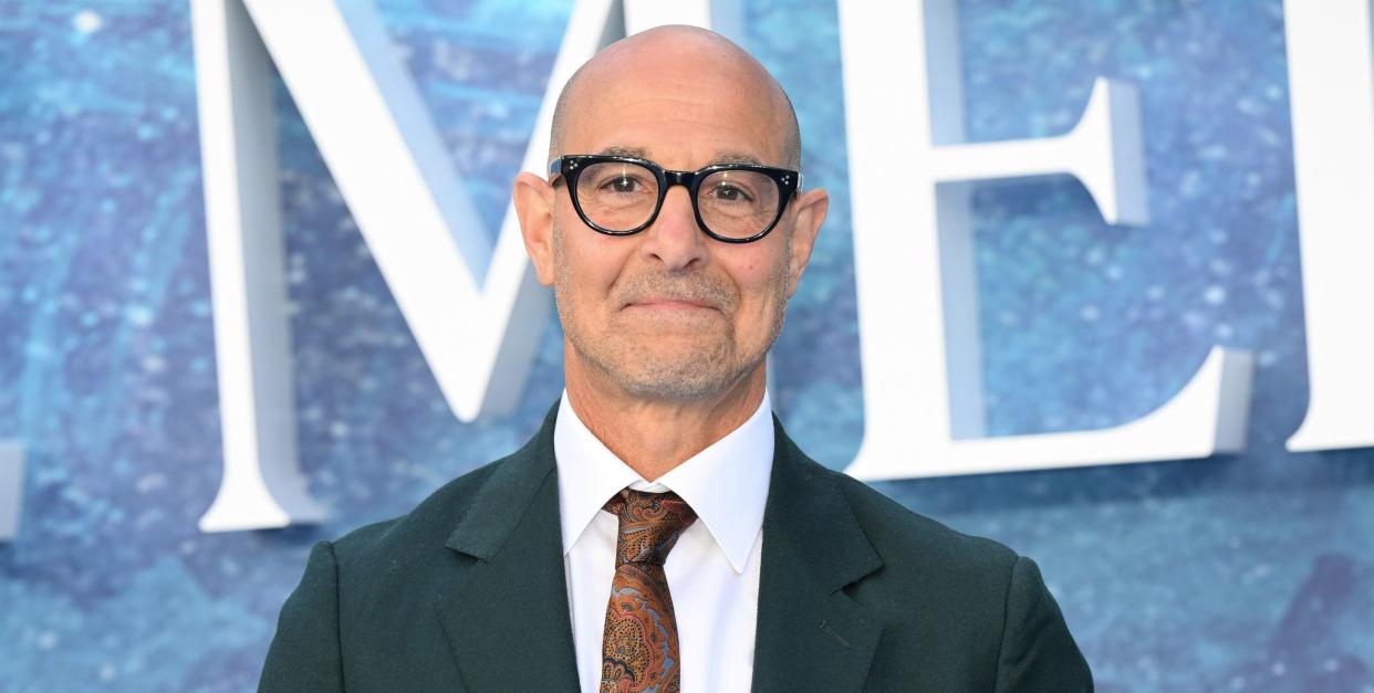 stanley tucci wearing a suit with a brown and orange paisley print tie and glasses on the red carpet at the little mermaid uk premiere