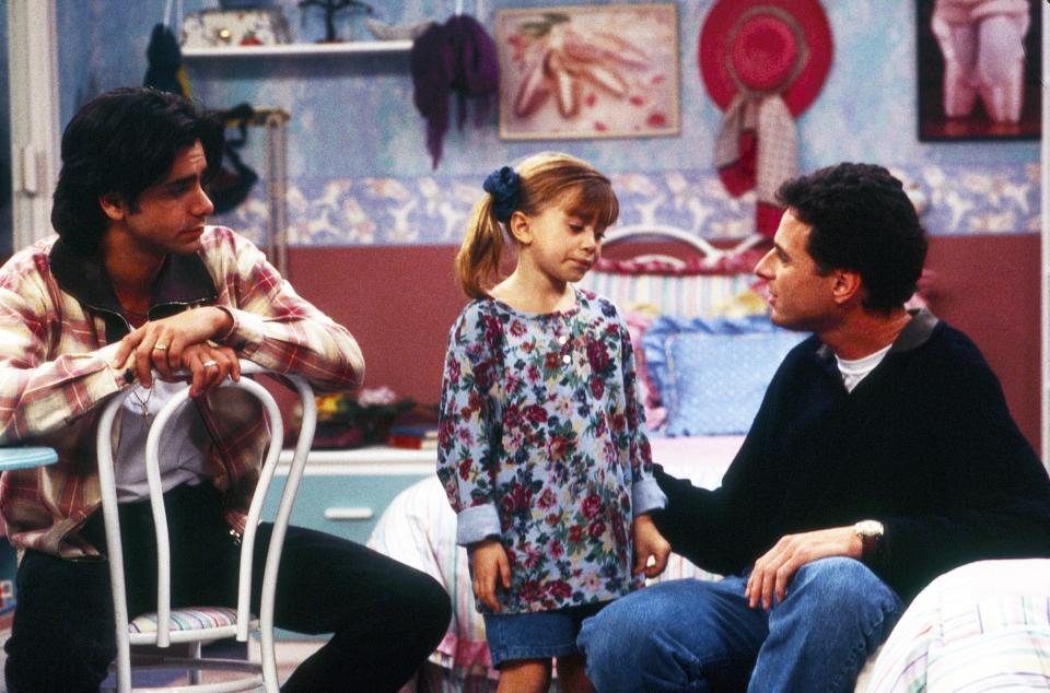 John Stamos, Mary-Kate/Ashley Olsen, and Bob Saget are in a scene from "Full House." The child wears a floral dress, and the men are casually dressed indoors
