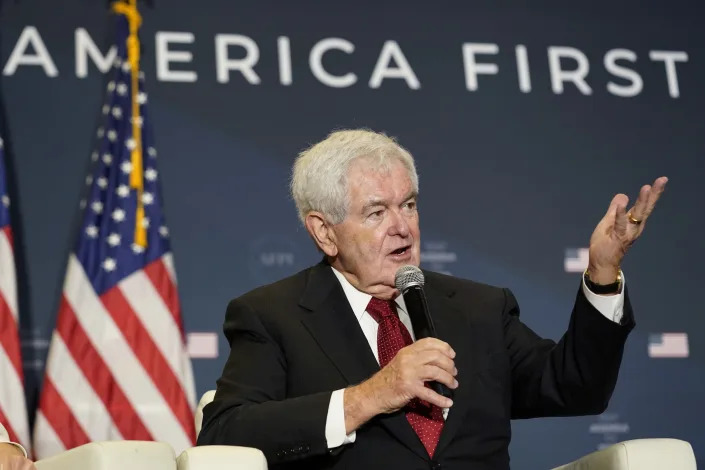 Former House Speaker Newt Gingrich sits onstage and speaks into a microphone at an America First Policy Institute summit in Washington.